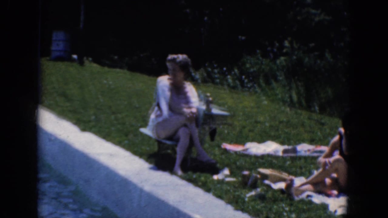 Adults relaxing on a sunny lawn beside a paved edge, with women seated on towels and a chair.