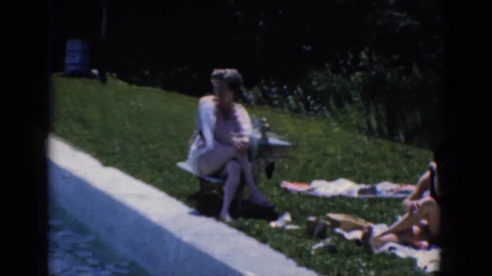 Candid 8mm frame of adults in swimsuits relaxing on grass near a paved edge during a sunny outdoor gathering.