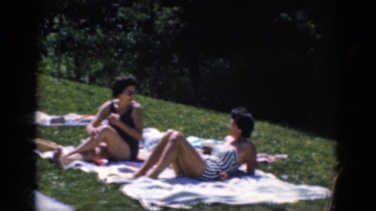 Two women in swimsuits recline and sit on towels spread across a grassy lawn.