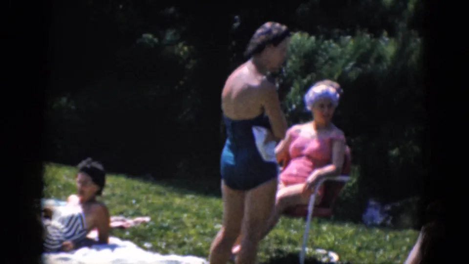 Adults gather informally on a lawn, including a standing woman in a swimsuit, a seated woman in a chair, and another person on a towel.