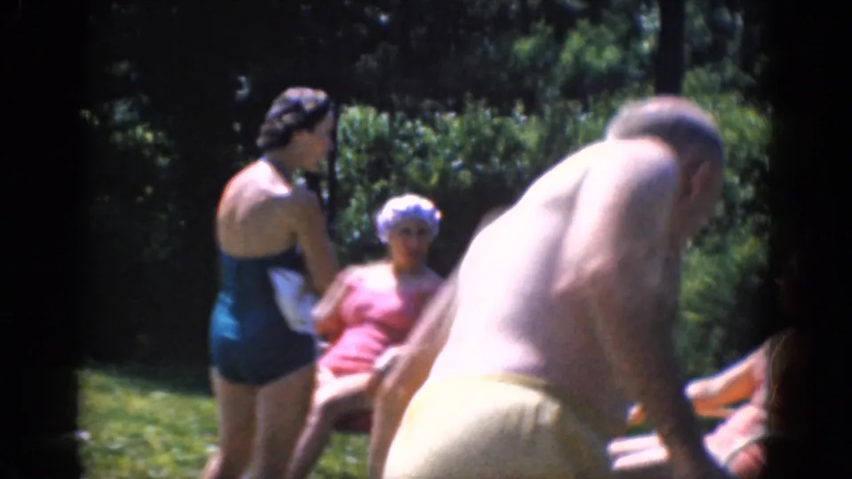 A man in light swim trunks walks close to the camera while women in swimsuits sit and stand on the lawn behind him.