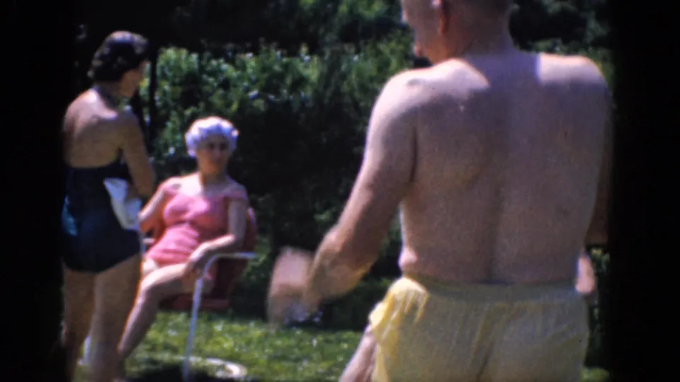 1968 8mm home-movie frame showing adults in swimsuits on a lawn, including a seated woman in pink and a standing woman in a dark one-piece.