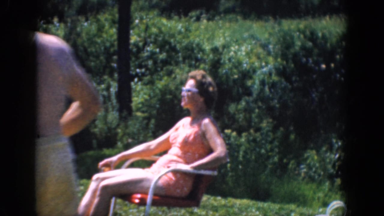 A woman in sunglasses sits in a lawn chair on grass with hedges behind her.