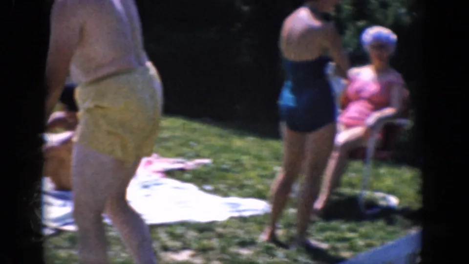 1968 8mm home-movie frame with a man in yellow trunks and women in bathing suits near towels on a sunny lawn.