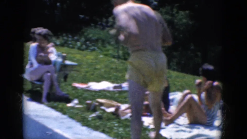 1968 8mm frame showing adults relaxing on a backyard lawn, with towels, chairs, hedges and a man in yellow swim trunks.