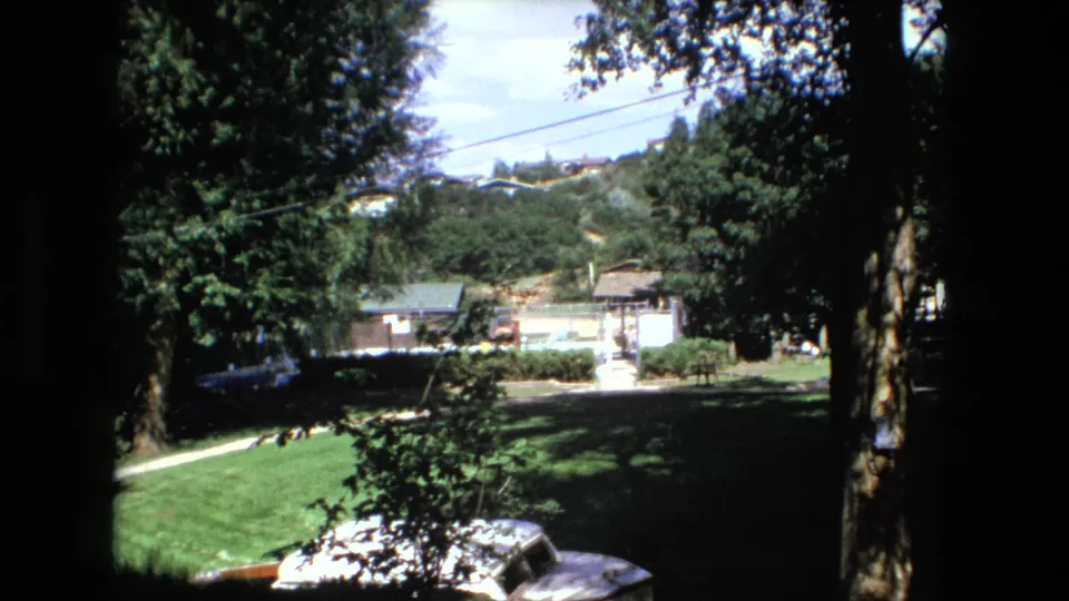 Fort Wayne, Indiana yard scene on 8mm film, showing a quiet lawn framed by dark tree trunks, with a small house and overhead power lines beyond.