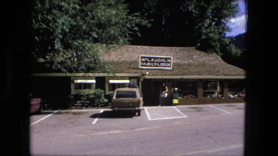 1976 Fort Wayne motel exterior with McLaughlin Family Lodge sign, brown station wagon, parking spaces, and woman by the doorway.