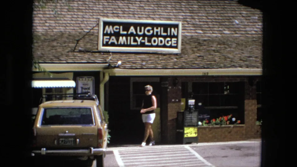 Closer 1976 view of McLaughlin Family Lodge with sign, shingled roof, parked station wagon, doorway, and woman in black top and white shorts.