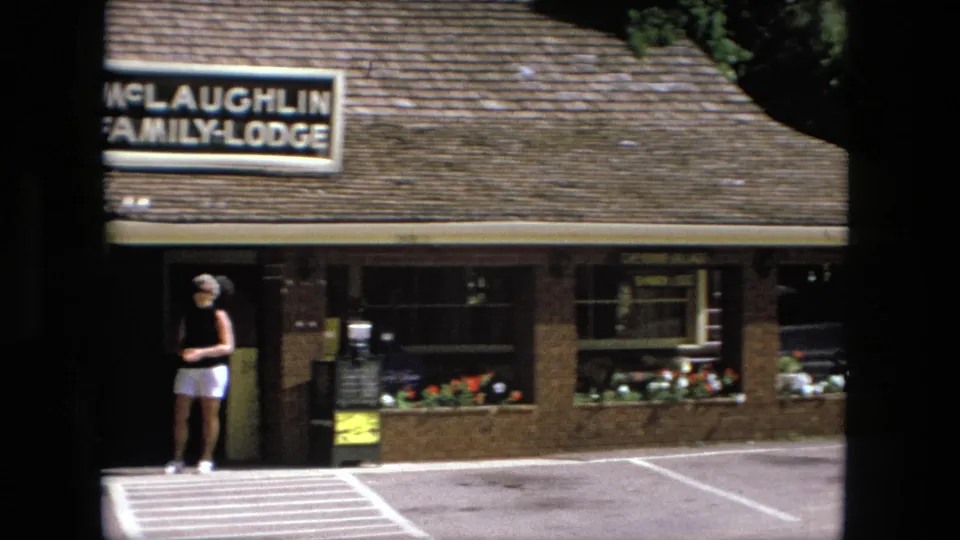1976 Fort Wayne motel exterior with the McLaughlin Family Lodge sign partly visible, woman at the doorway, front windows, and parking lot.