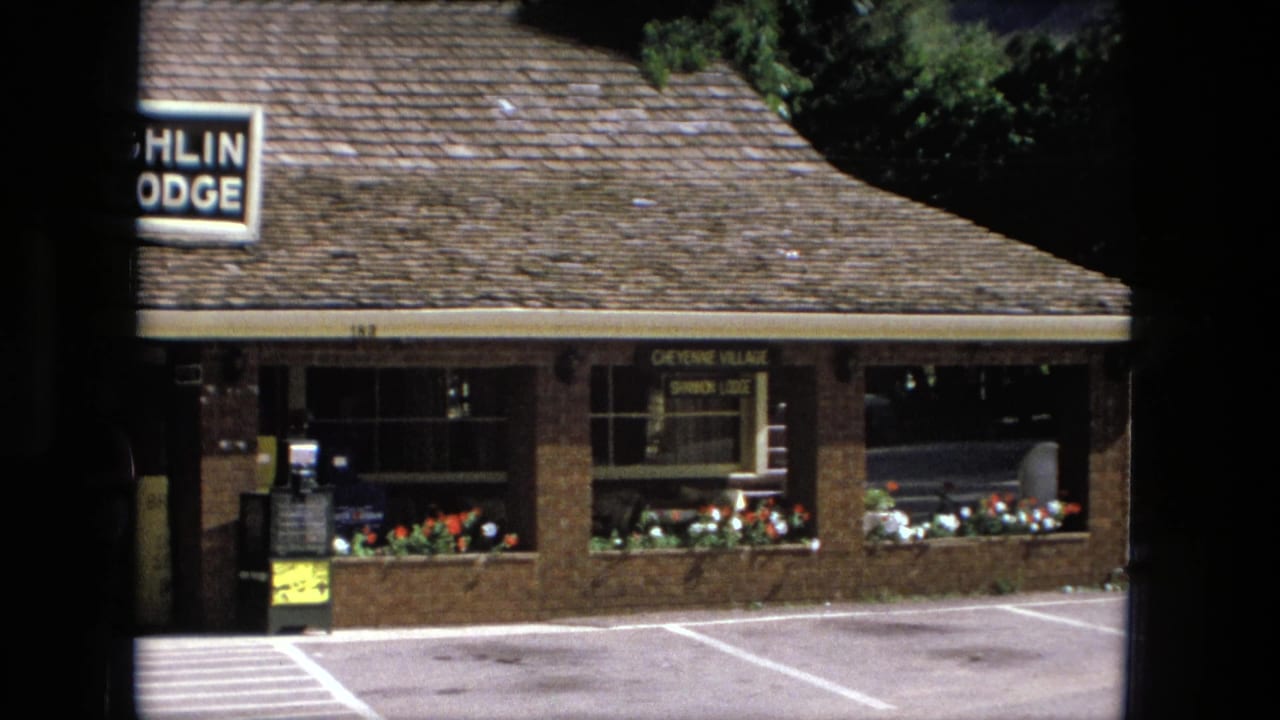 Brick low-rise lodge building with shingled roof, flower boxes, parking lines, and partial sign at left.