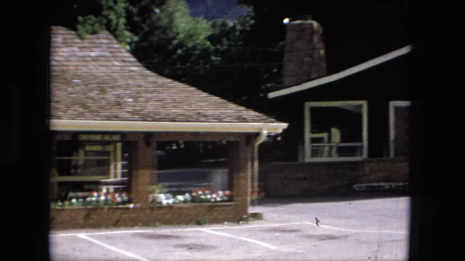 1976 Fort Wayne home-movie frame continuing across the brick frontage toward a dark single-storey house beside the parking area.