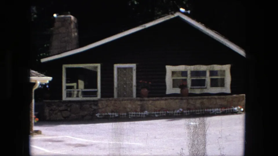 1976 Fort Wayne frame showing a dark single-storey house with stone chimney, front windows, low stone wall, and paved parking area.