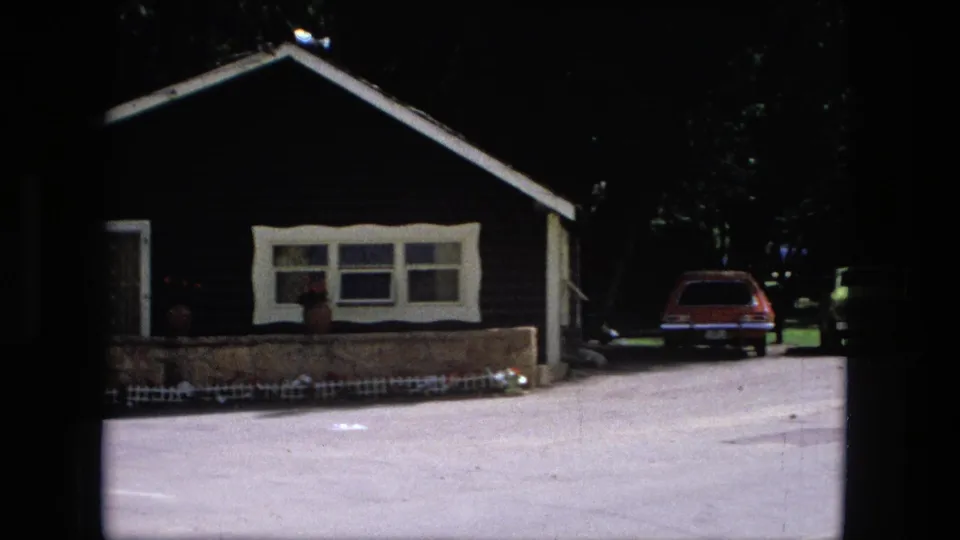 1976 Fort Wayne 8mm frame with a dark single-storey house, paved lot, trees, red station wagon, and lime green car.