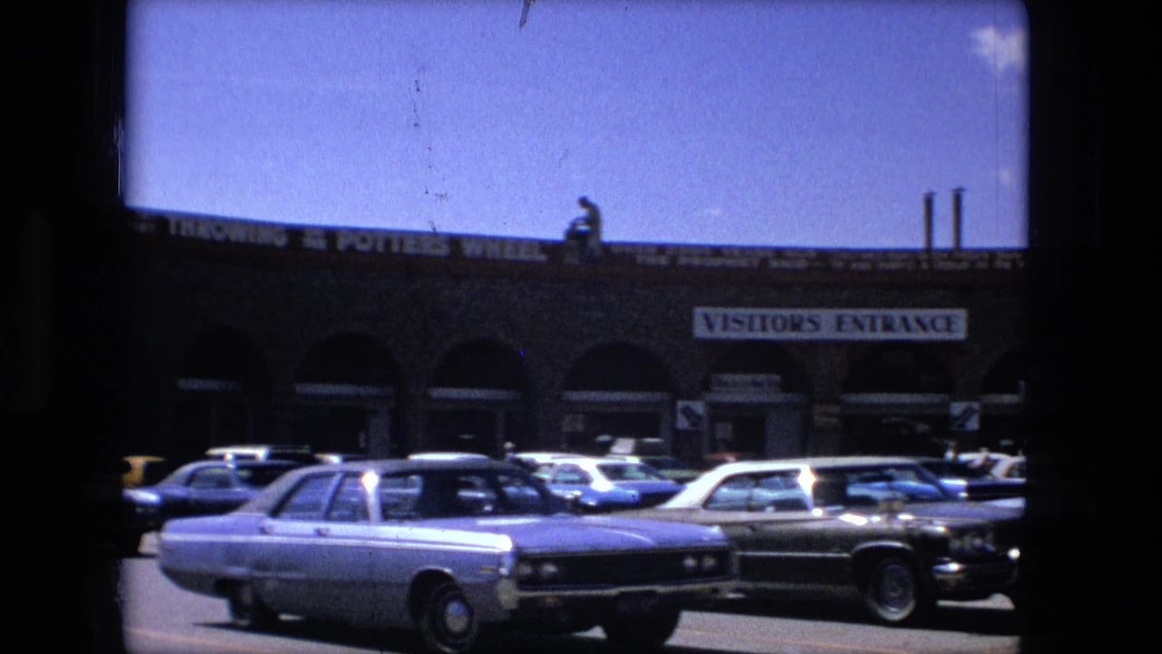 Tan stone shopping center with visitor entrance sign and parked 1970s cars in front.
