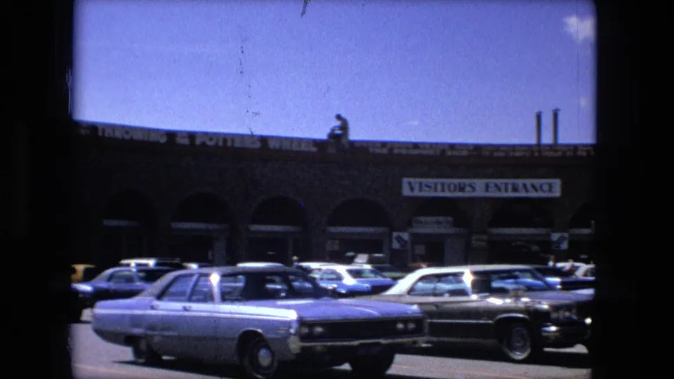 1976 Fort Wayne home-movie frame showing a tan stone commercial building, visitor entrance sign, and parked mid-1970s cars under a blue sky.