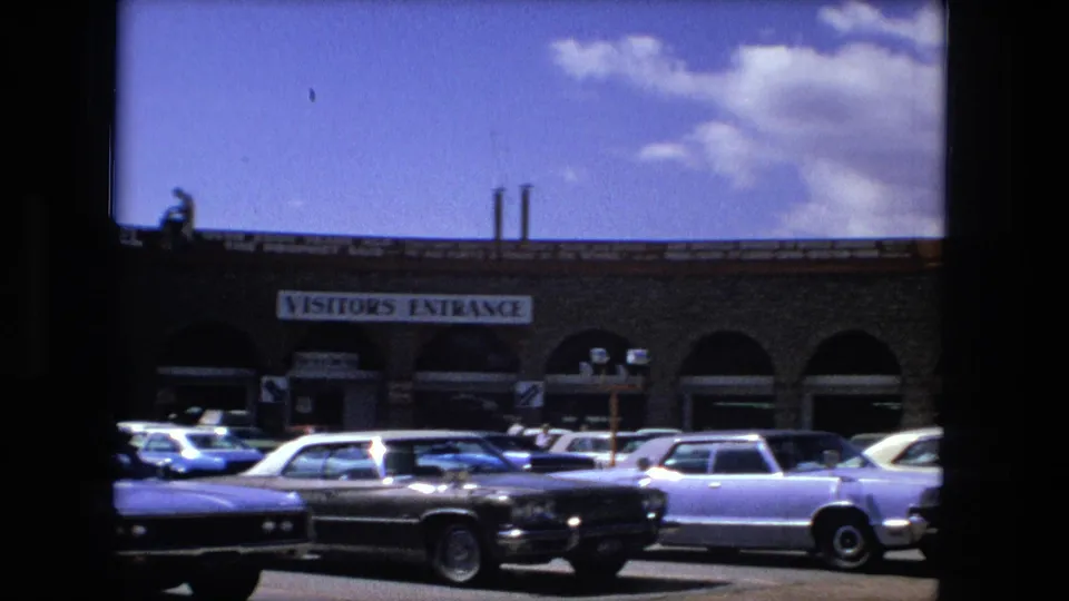 Fort Wayne 1976 frame with parked sedans, arched storefront openings, visitor entrance signage, and a small figure along the roofline.