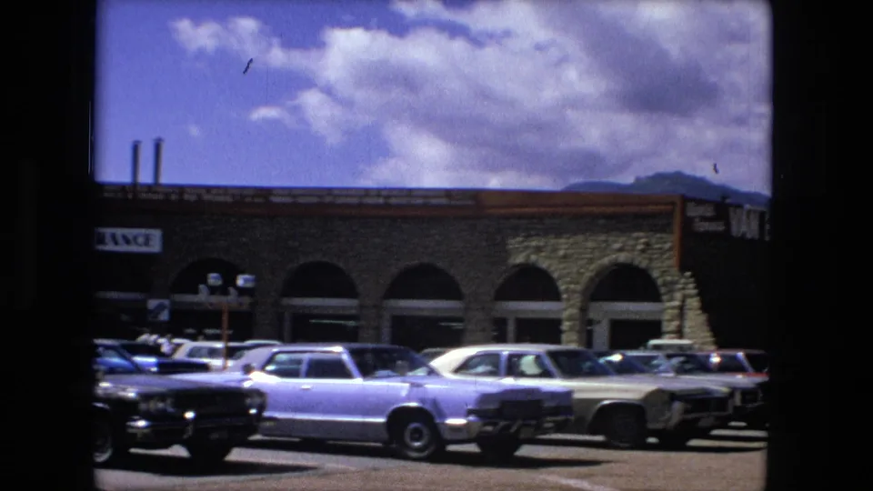 1976 Fort Wayne frame showing the shopping-center corner, arched openings, parked sedans, partial signage, and distant hills beneath clouds.