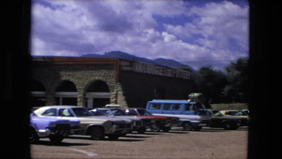 Fort Wayne 1976 home-movie frame with parked cars and a blue van outside a tan stone shopping center, with hills and clouds in the distance.