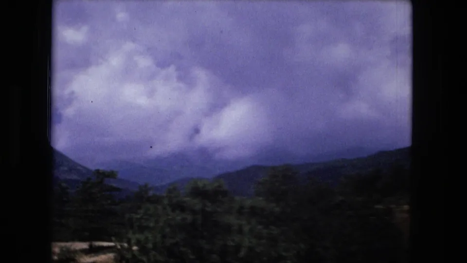 1970s 8mm frame of layered mountain ridges under a cloudy sky, with trees below and dark rounded film edges.