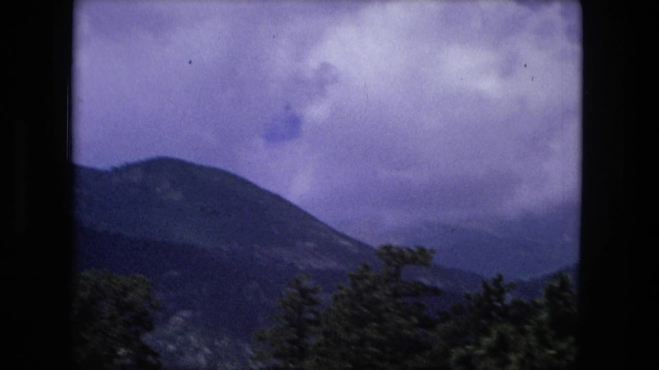 A large dark mountain slope rises behind evergreen trees in an aged 8mm home-movie frame cataloged to 1976.