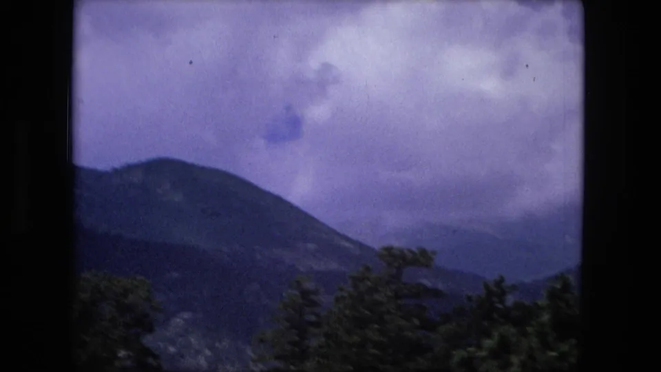 A large dark mountain slope rises behind evergreen trees in an aged 8mm home-movie frame cataloged to 1976.