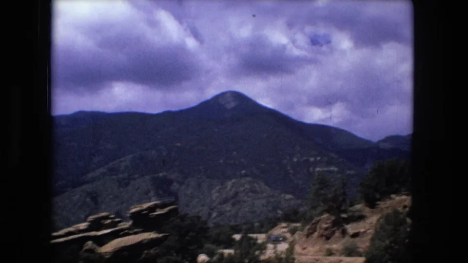A rocky overlook faces tree-covered mountains beneath heavy clouds, with visible aged-film grain and purple-blue tint.