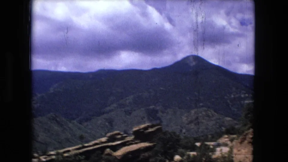 1970s 8mm frame of distant mountain ridges and a cloudy sky seen from a high overlook, with dark gate edges visible.