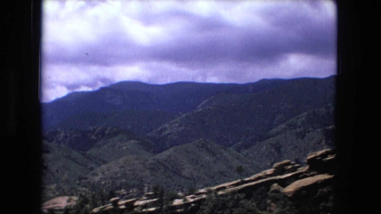 1970s 8mm mountain landscape view from an elevated position, showing ridges, green slopes, and purple-blue cloud cover.