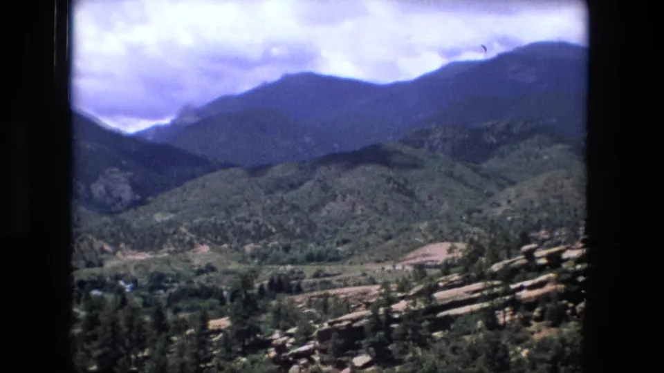 1970s 8mm scenic overlook view with rocky ledges, green mountain slopes, a valley, and clouded sky.