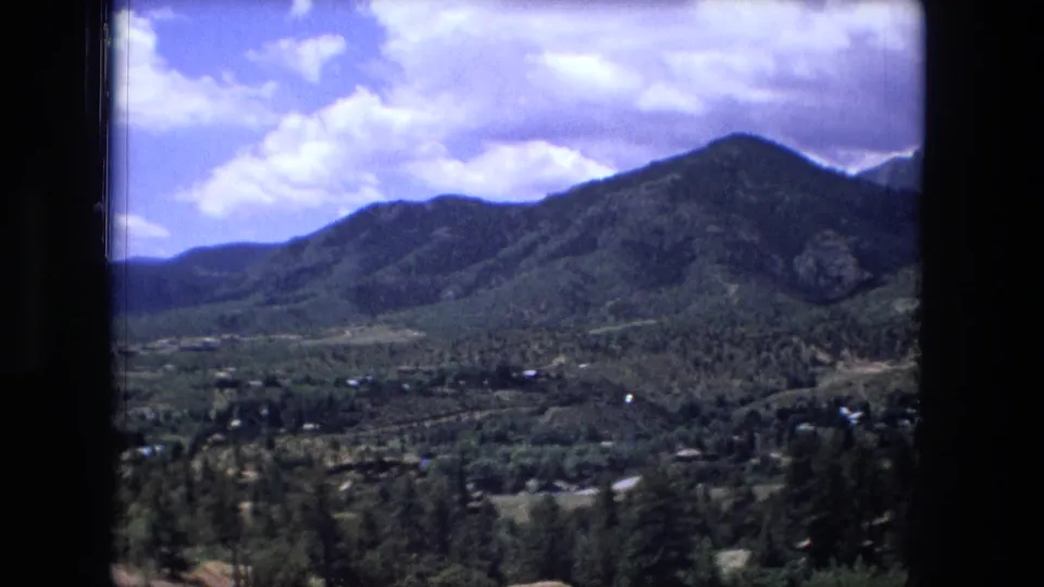 1976 8mm home-movie frame showing a central wooded mountain slope with a settled valley and scattered trees below.