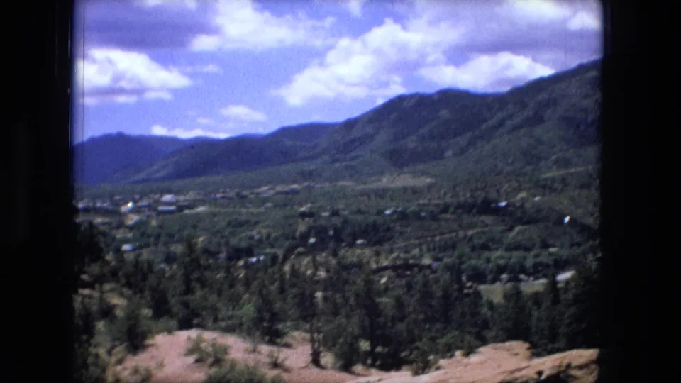 1976 8mm frame showing the pan opening onto a wider valley with green hillsides, trees, small buildings, and clouds.