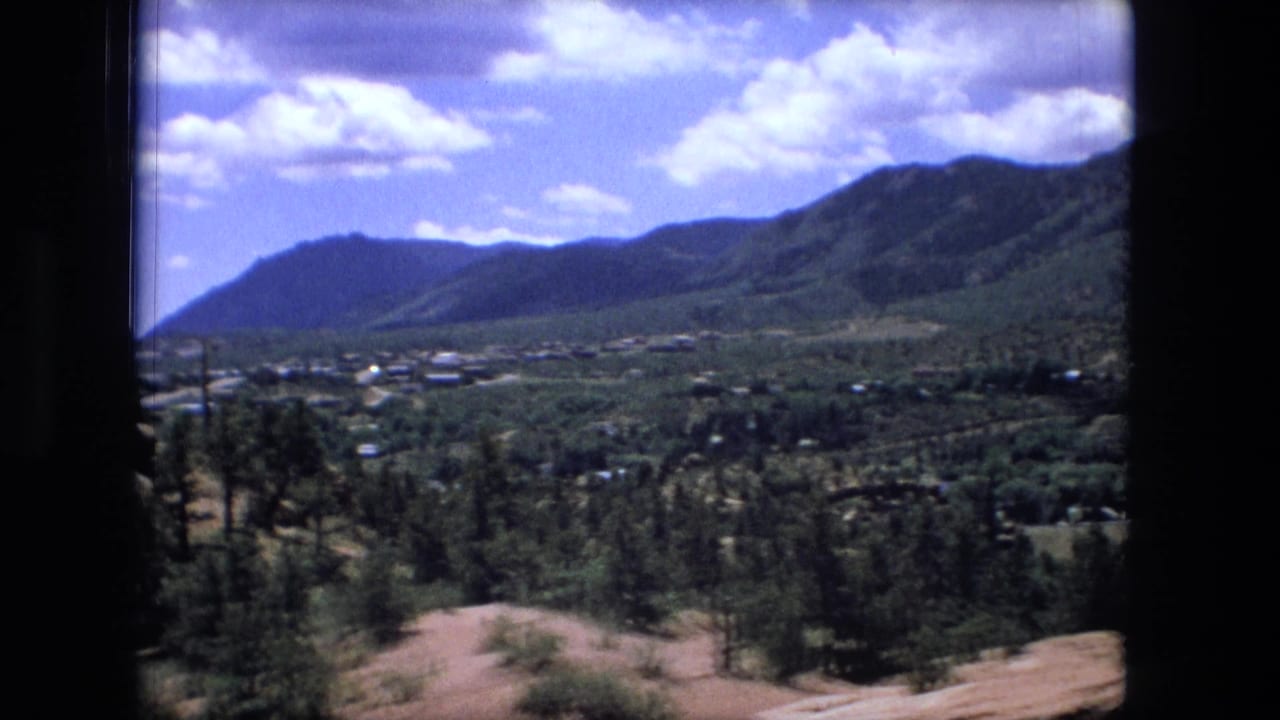 Mountain valley with scattered buildings, trees, rocky foreground, and blue sky with clouds.