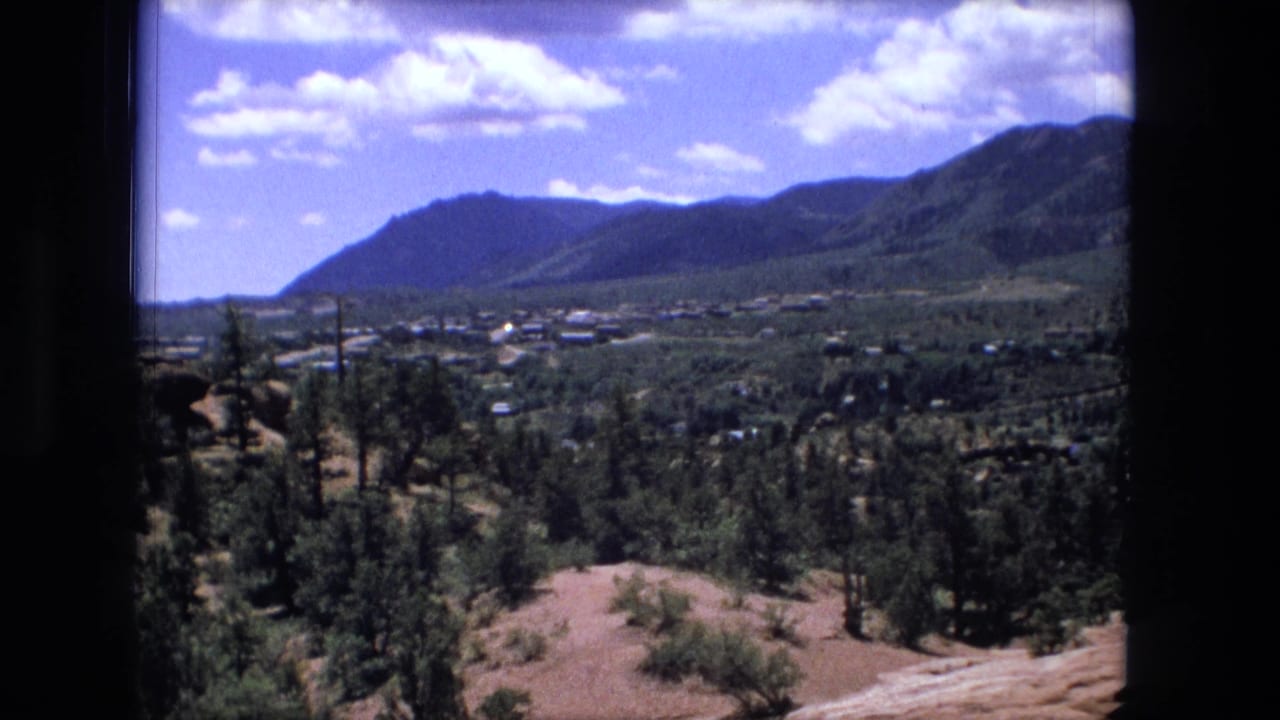 Wide valley landscape with forested slopes, distant buildings, shrubs, and bright clouded sky.