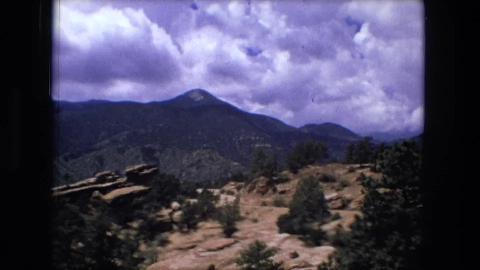 1976 8mm frame cataloged to Fort Wayne, Indiana, showing rocky terrain and trees before dark mountain ridges under large clouds.