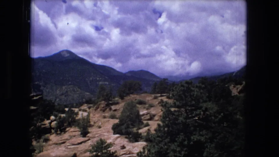 1976 8mm frame cataloged to Fort Wayne, Indiana, showing shrubs and rock outcrops facing mountain ridges beneath a cloud-filled sky.