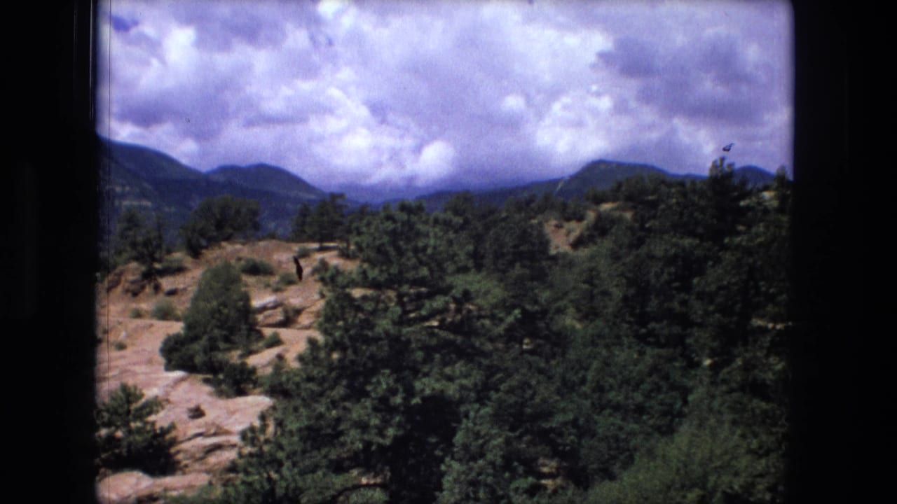 8mm frame from the labeled 1976 clip showing a pine-covered slope, rocky terrain, distant ridges, and dramatic clouds.