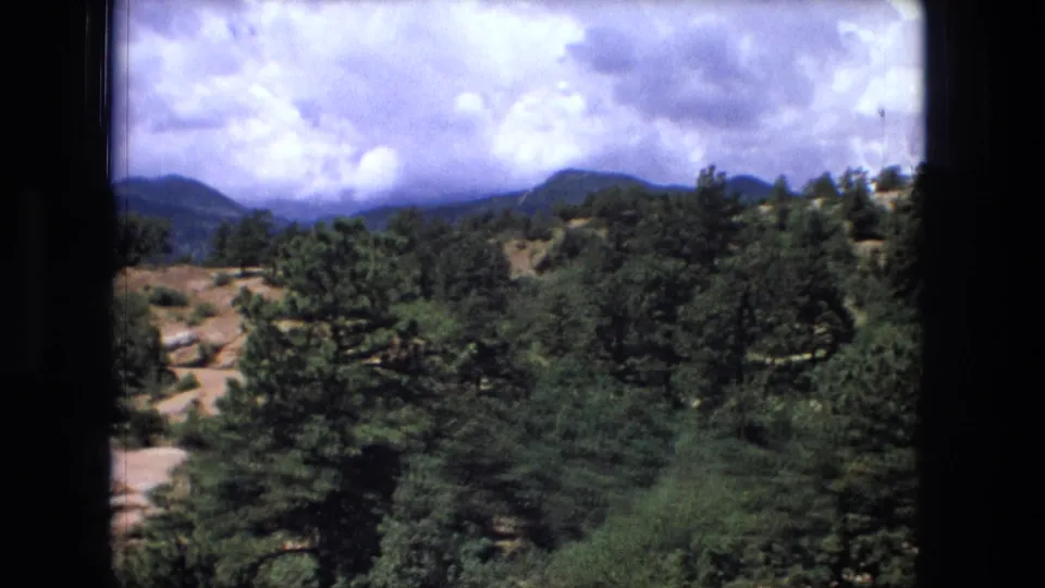 Vintage 8mm landscape view with dense foreground trees, rolling hills, distant mountains, and a bright clouded sky.