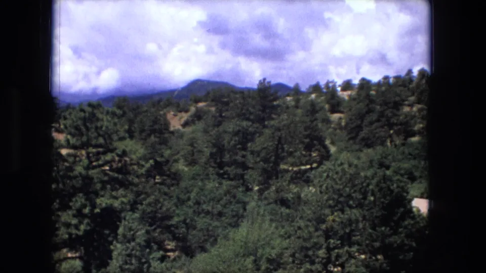 1976 8mm frame showing a wooded hillside, darker green trees, distant ridges, and bright clouds across the sky.