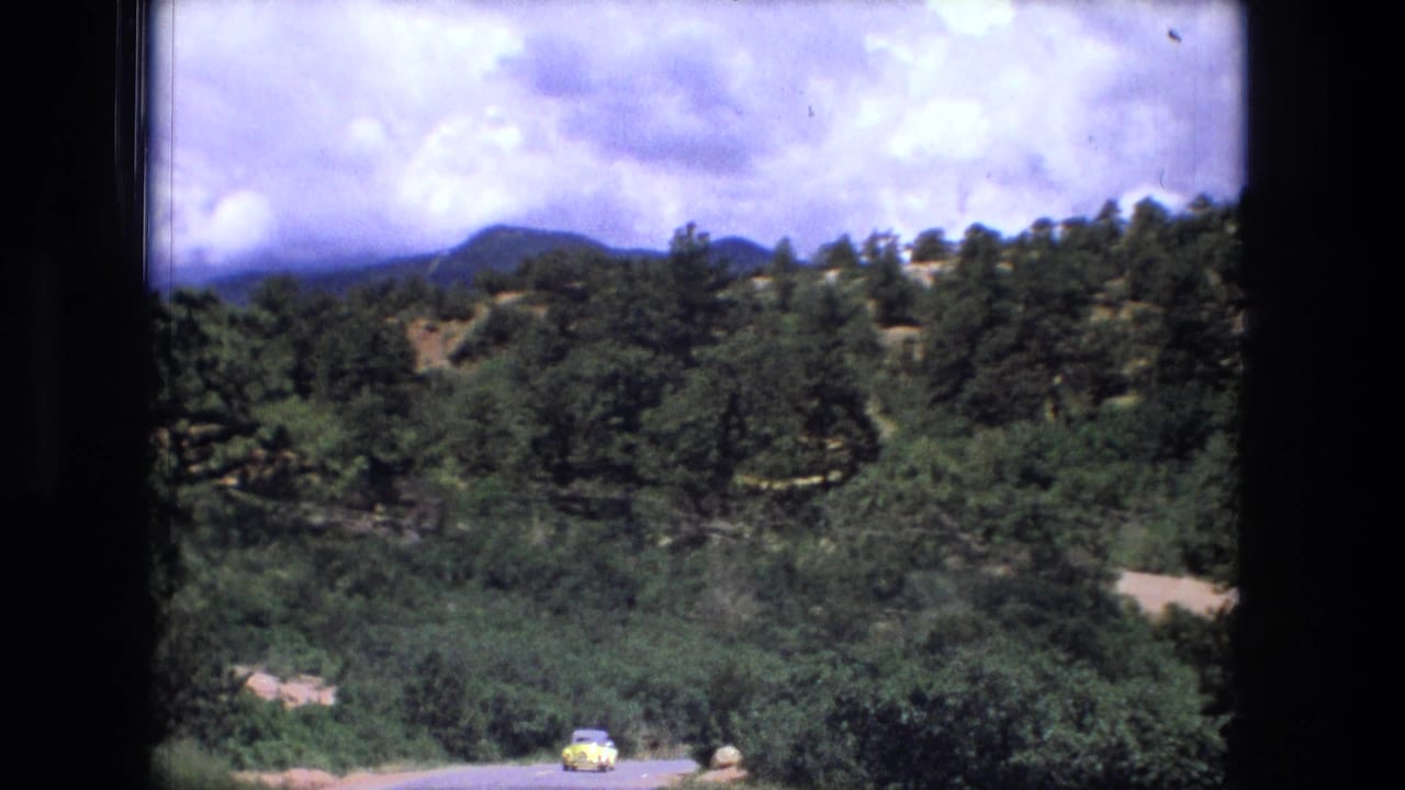 Rocky green valley with a road and small yellow car under a cloudy blue sky.