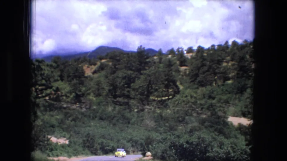 1976 8mm frame of a green valley road with a small yellow car, rocky hills, trees, and bright clouds, as cataloged to Fort Wayne, Indiana.