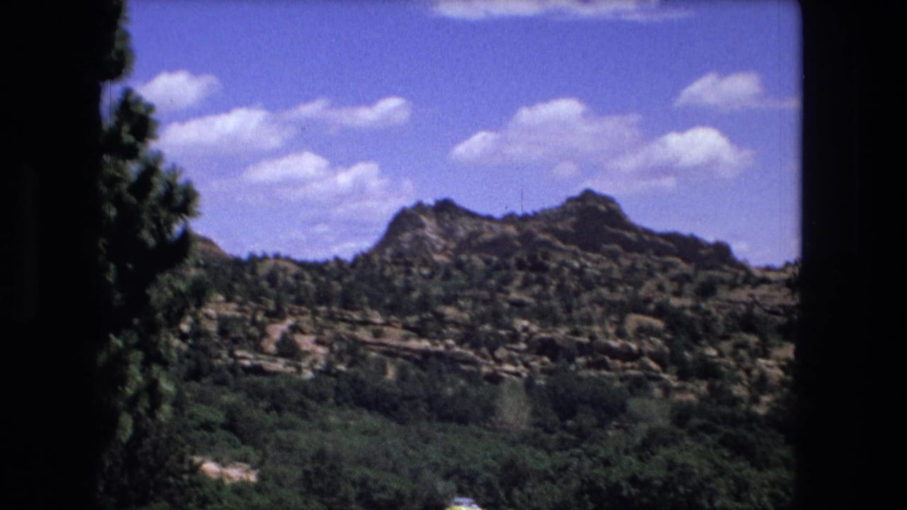1976 8mm frame showing a rocky ridge above dense green vegetation and a blue sky, with dark home-movie frame borders.