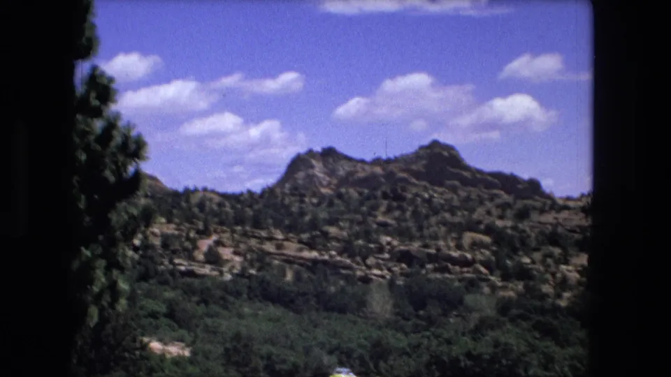 1976 8mm frame showing a rocky ridge above dense green vegetation and a blue sky, with dark home-movie frame borders.
