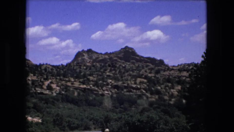 1976 8mm frame of a rocky, shrub-covered hillside and valley vegetation beneath scattered clouds, with soft color film grain.