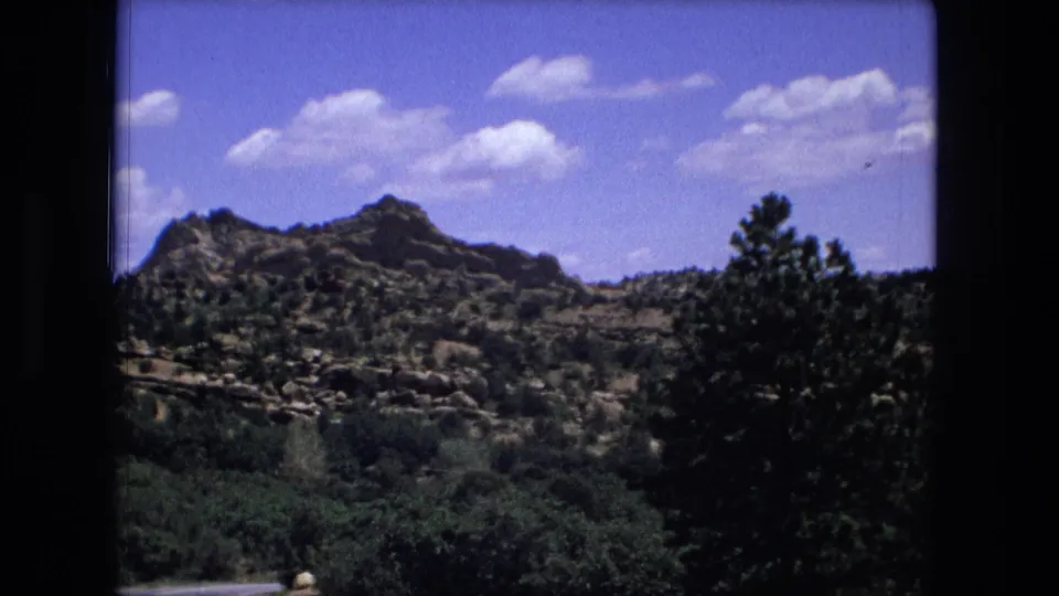 1976 8mm frame of a rocky hillside and green valley brush, partly framed by a dark tree silhouette and film borders.