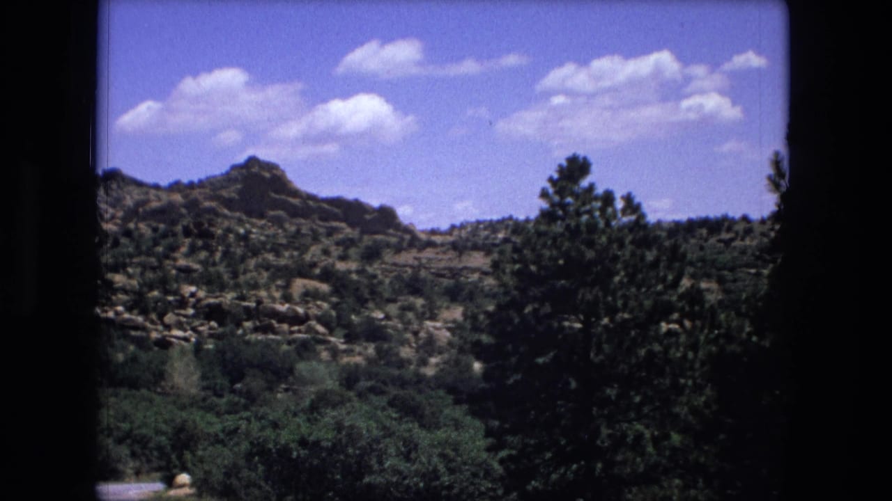 Wooded rocky hills under a blue sky with scattered clouds and dark film borders.
