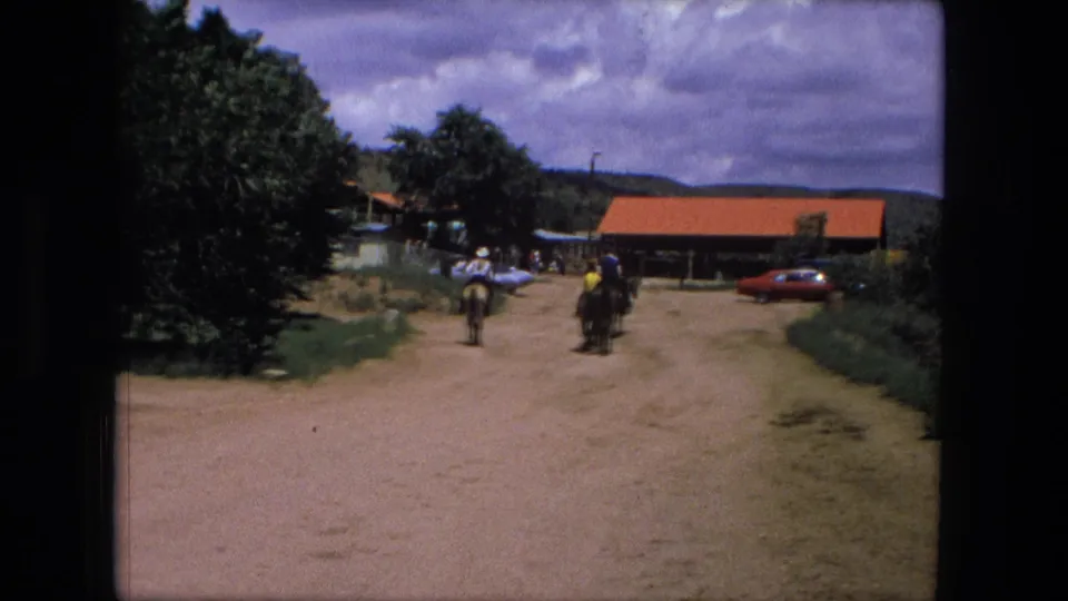 People on horseback ride along a dirt road toward rustic red-roofed buildings, with parked cars nearby in 1976 home-movie footage.