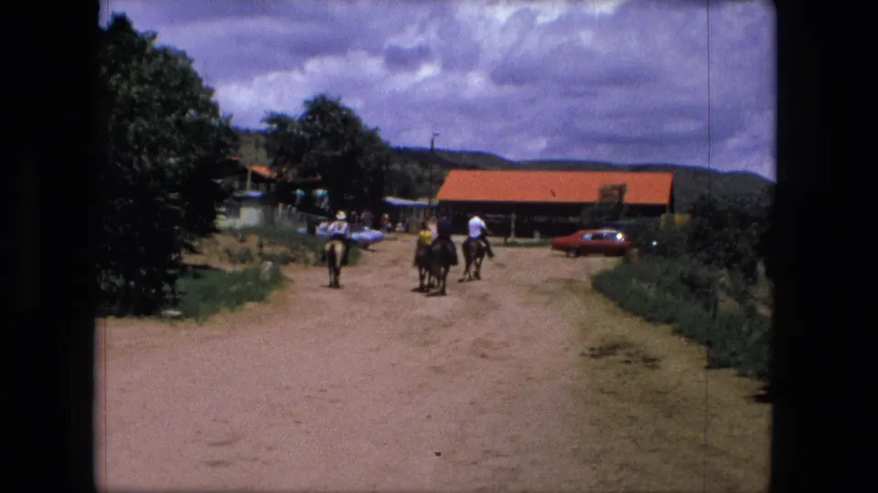In a 1976 8mm frame, several riders on horseback pass along a dirt road near red-roofed buildings, trees, and vintage cars.