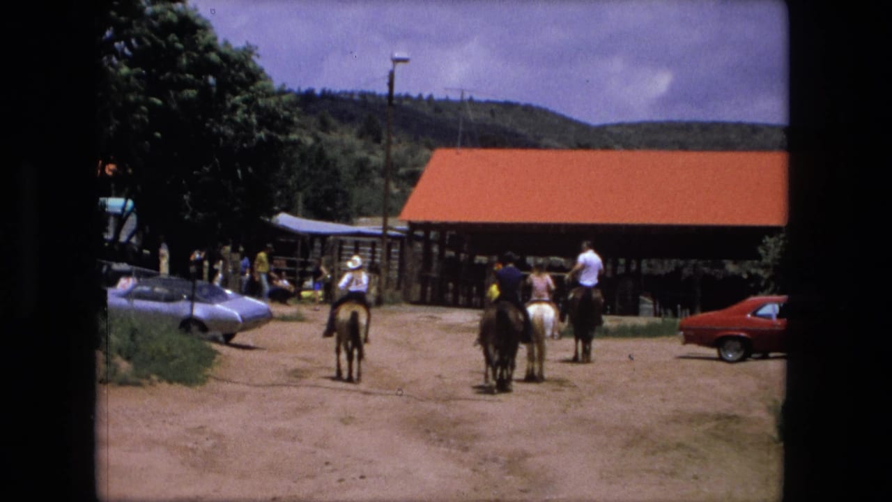 Several people ride horses across a dusty ranch yard near parked cars and a red-roofed barn.