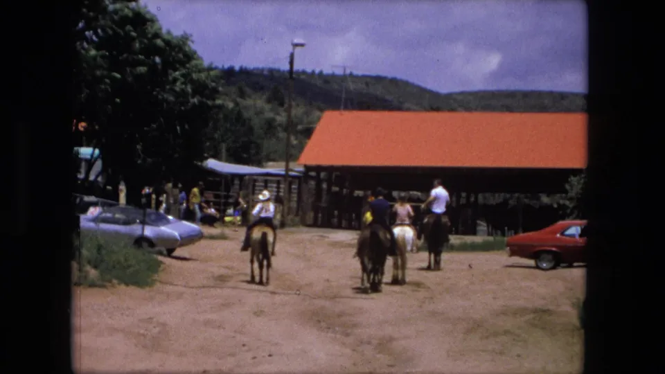 1976 Fort Wayne home-movie frame showing horseback riders moving toward a red-roofed barn with vintage cars nearby.