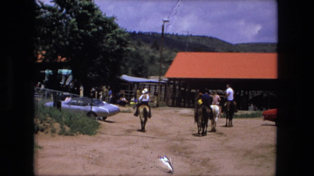 1976 Fort Wayne ranch scene with riders on horseback passing between parked cars and a red-roofed barn.